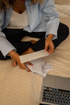 Individual sitting cross-legged examining papers near laptop indoors.